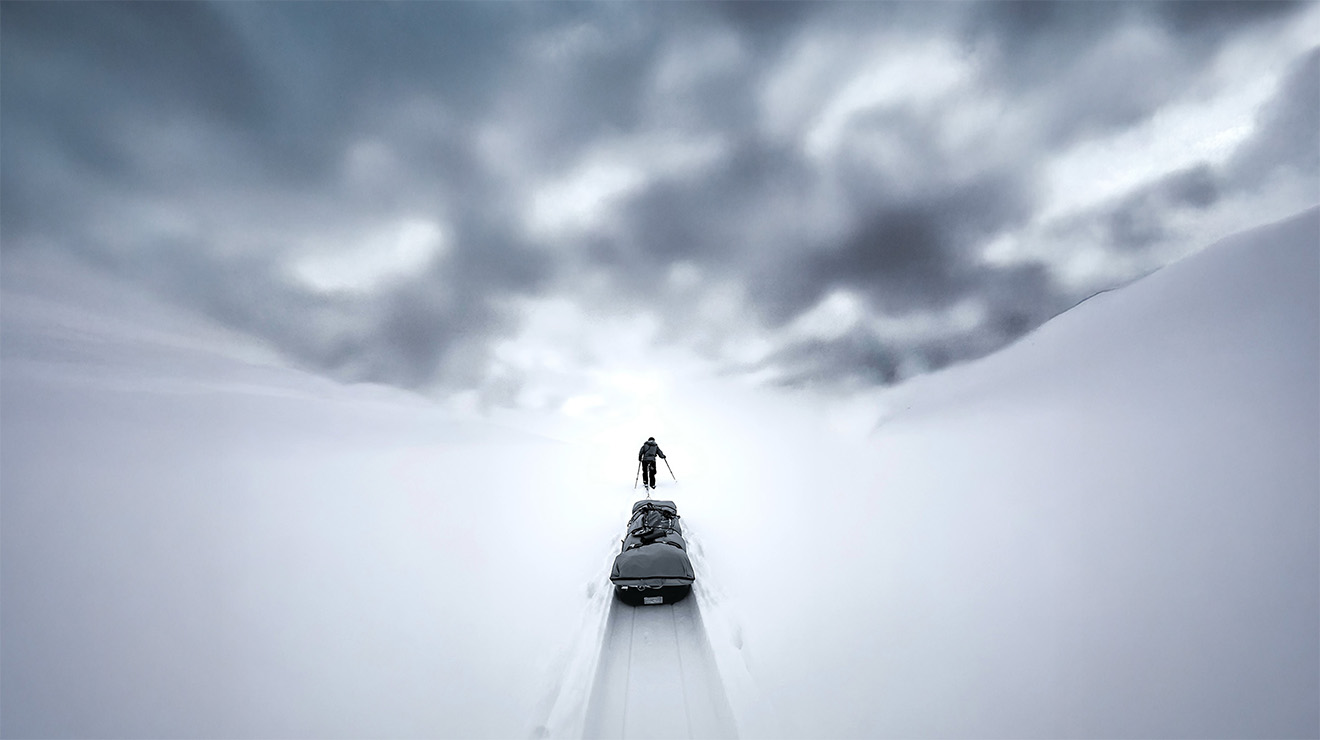A lone figure hauling a sled across the Greenland ice sheet under a heavy sky