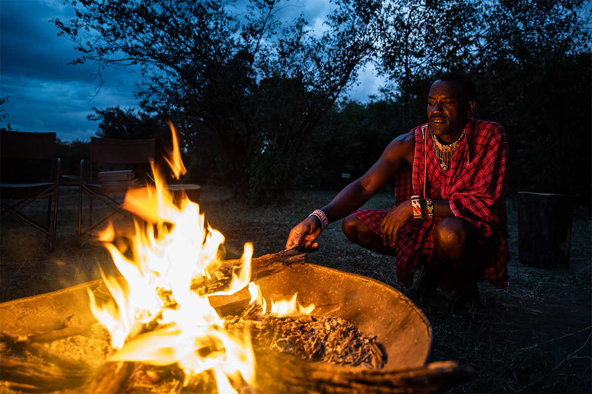 A Maasai man in a red shuka tending the campfire at dusk